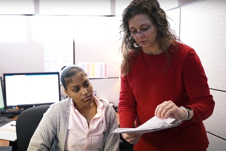 a woman in a red shirt goes over paperwork with an intern sitting at her computer